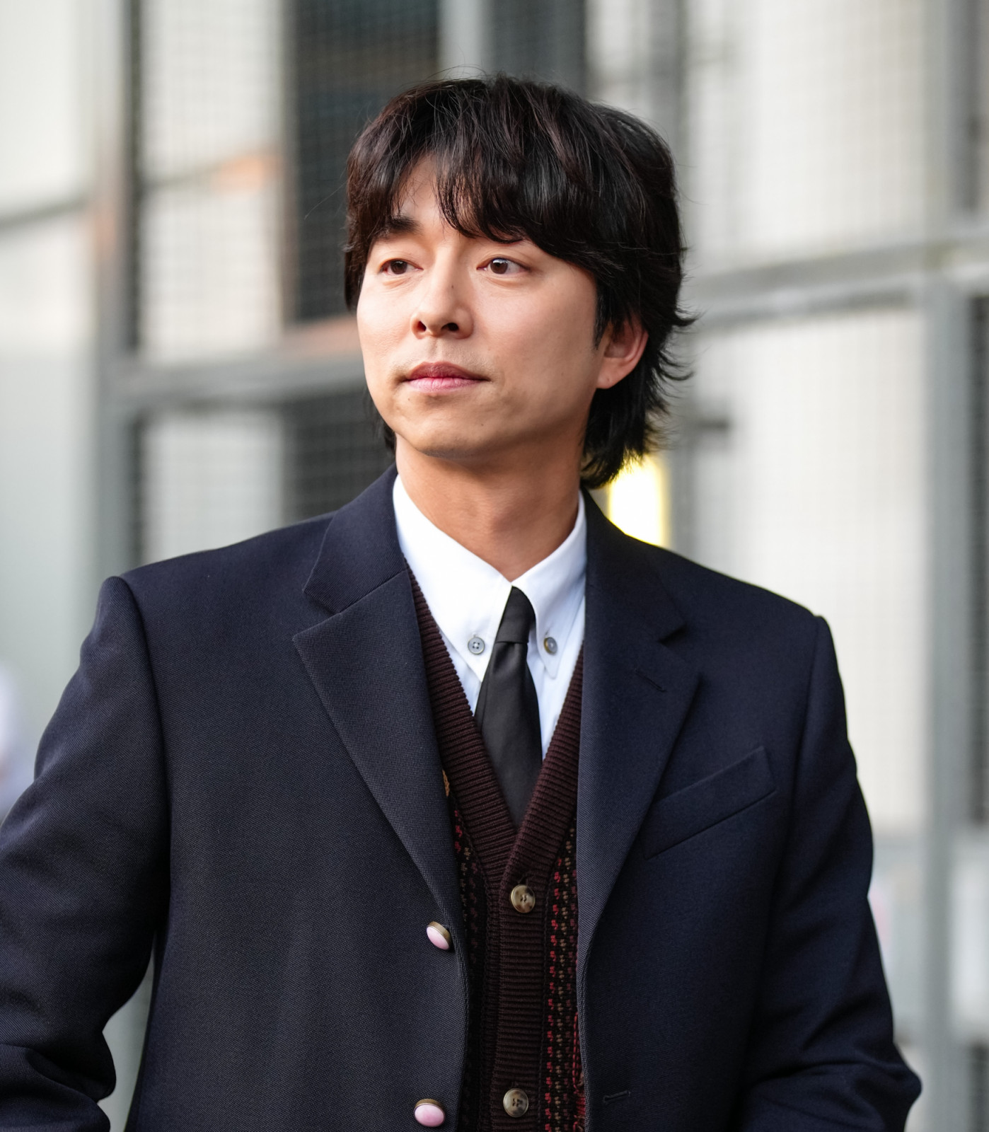 PARIS, FRANCE - JUNE 24: Gong Yoo wears a dark navy blazer with structured shoulders and a single-breasted design, featuring metallic buttons. Underneath, a white collared shirt is visible, paired with a black tie. A dark cardigan with subtle texture is layered beneath the blazer. The hairstyle is medium-length and styled with a natural texture, outside Louis Vuitton, during Paris Fashion Week - Menswear Spring/Summer 2026, on June 24, 2025 in Paris, France (Photo by Edward Berthelot/Getty Images)