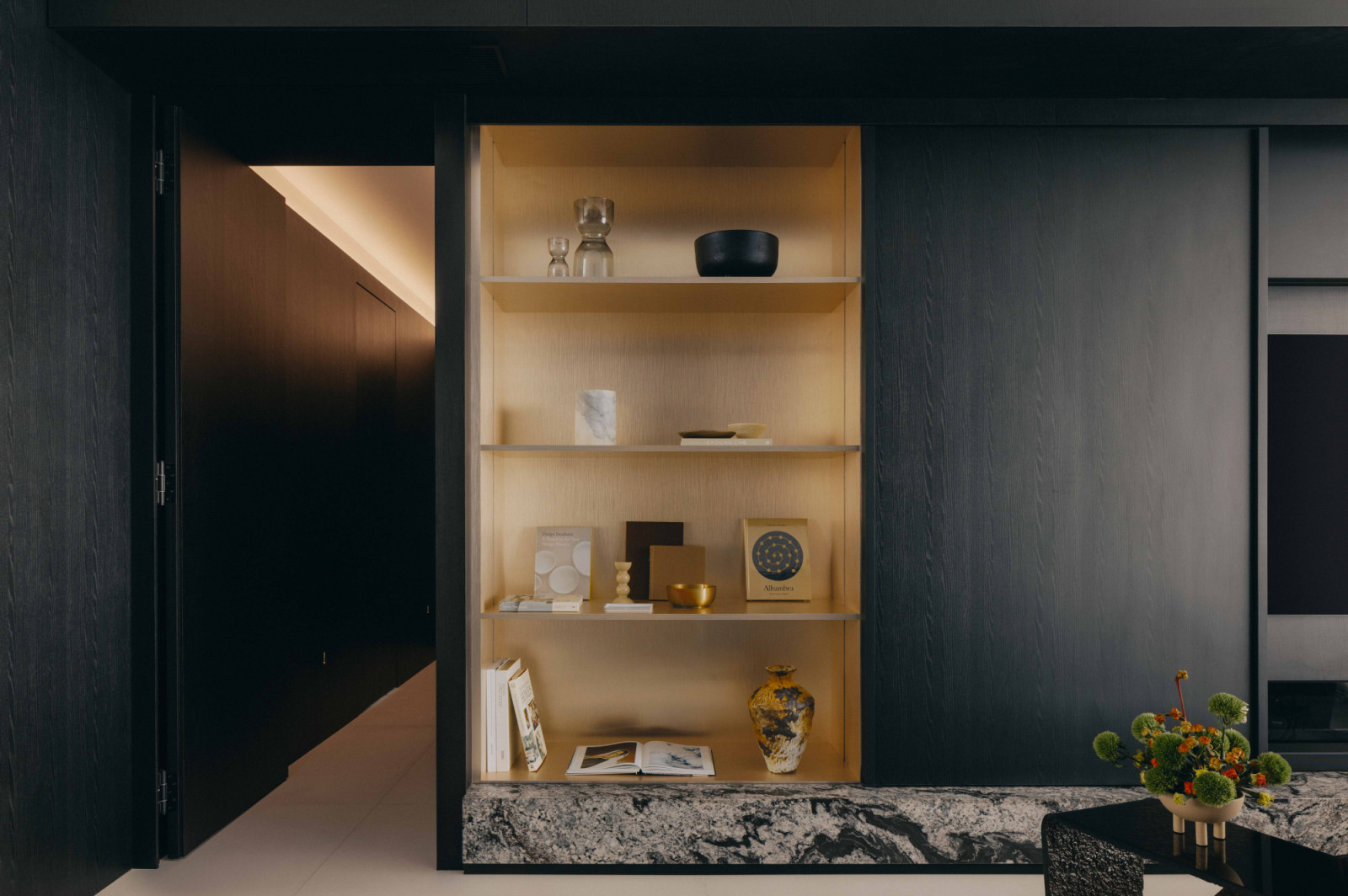 Display shelves in pale wood with softly lit niches punctuate the apartment’s dark carpentry