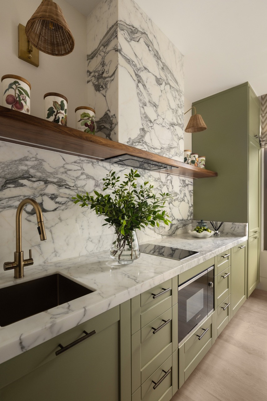 A secondary kitchen features sage green cabinetry paired with marble countertops and a full-height marble splashback beneath a timber display shelf