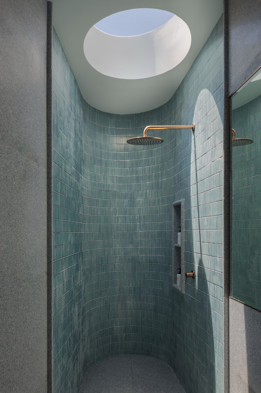 A shower alcove features vertically stacked teal mosaic tiles beneath a circular skylight with brass rainfall fixtures