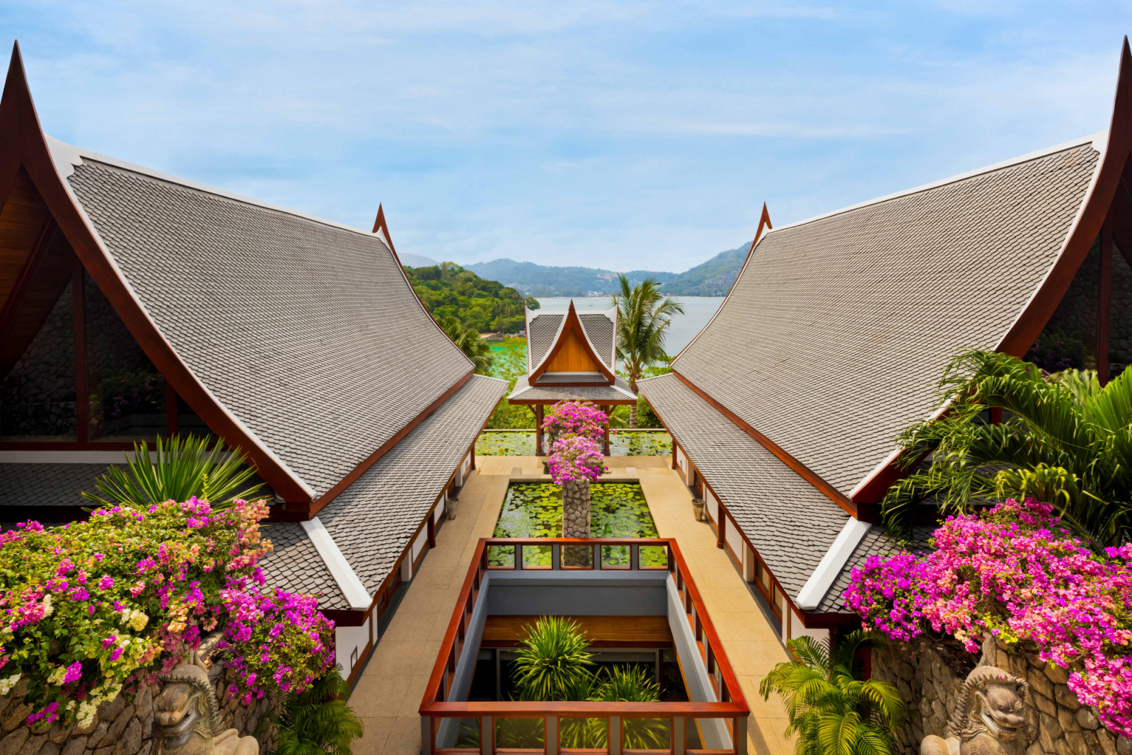 Overhead view of the sala roofs framing a central void that looks down to the lower level, with bougainvillea in full bloom softening the geometry inspired by the vernacular Thai architecture