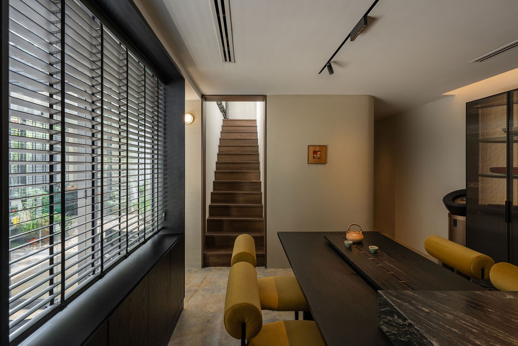 The walnut-clad staircase rises adjacent to the dining area, with yellow seating and grey-tinted glass panels defining the space
