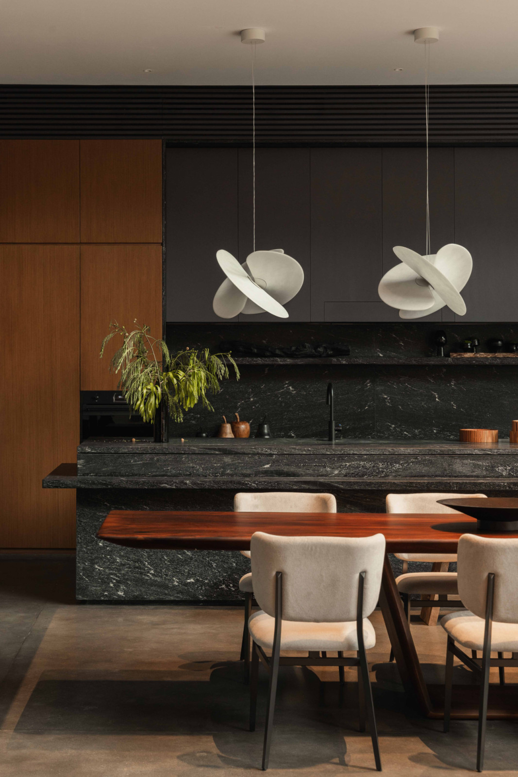 Light-coloured dining chairs and pendant lights contrast with the dark-finished kitchen beyond