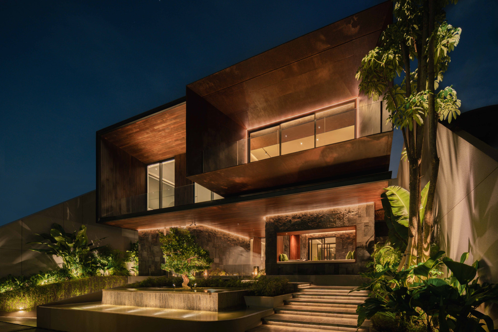 The house presents a corten steel facade in the evening, its cantilevered box volumes articulated via light and shadow