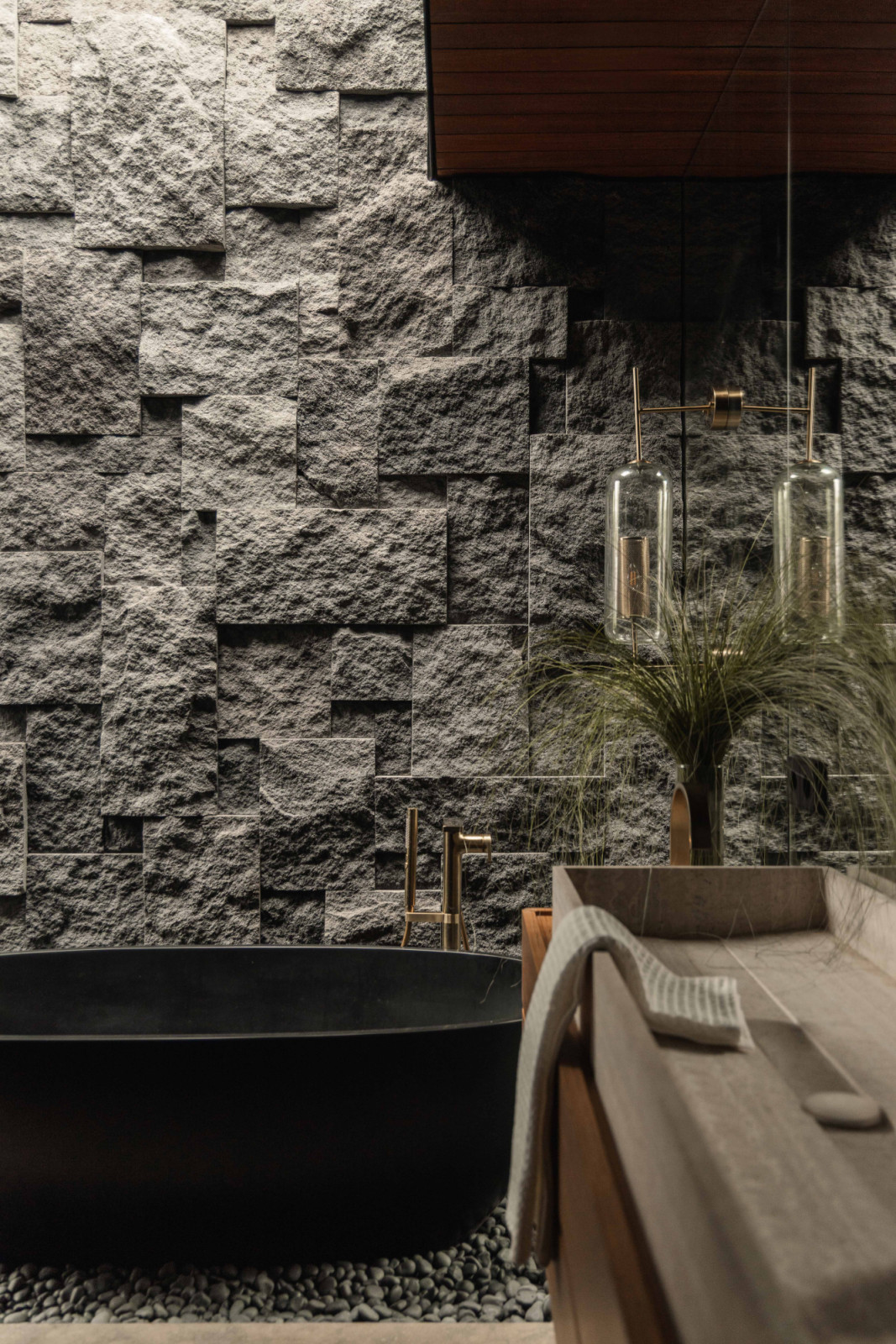 A freestanding bathtub is set against a natural stone wall with pronounced texture, lit by daylight from the skylight above