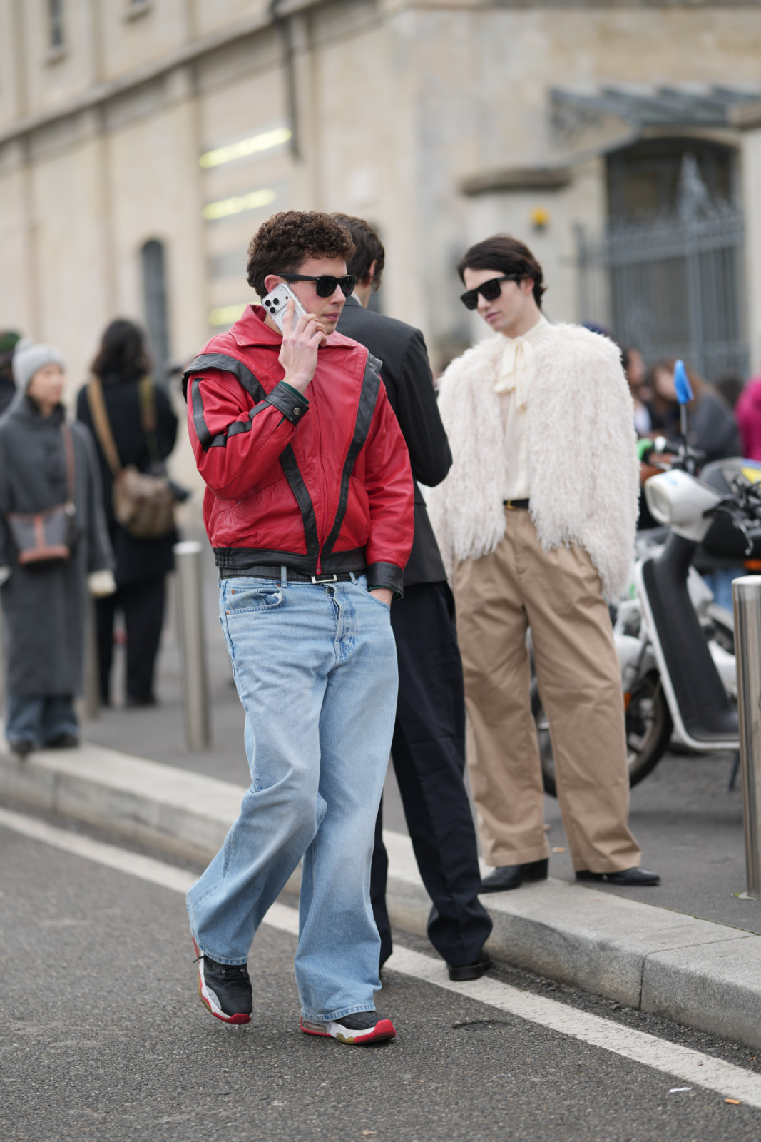 MILAN, ITALY - JANUARY 18: A guest wears short curly dark brown hair, black rectangular sunglasses, a red leather jacket with black leather trim and paneling, zip-front closure, stand collar and black elastic hem and cuff details, light blue faded denim jeans in a loose wide-leg cut with five-pocket styling and a visible black leather belt with a silver buckle, black and gray leather-and-mesh sneakers shoes with white midsoles and red rubber outsoles, outside Prada, during Milan Fashion Week - Menswear Fall