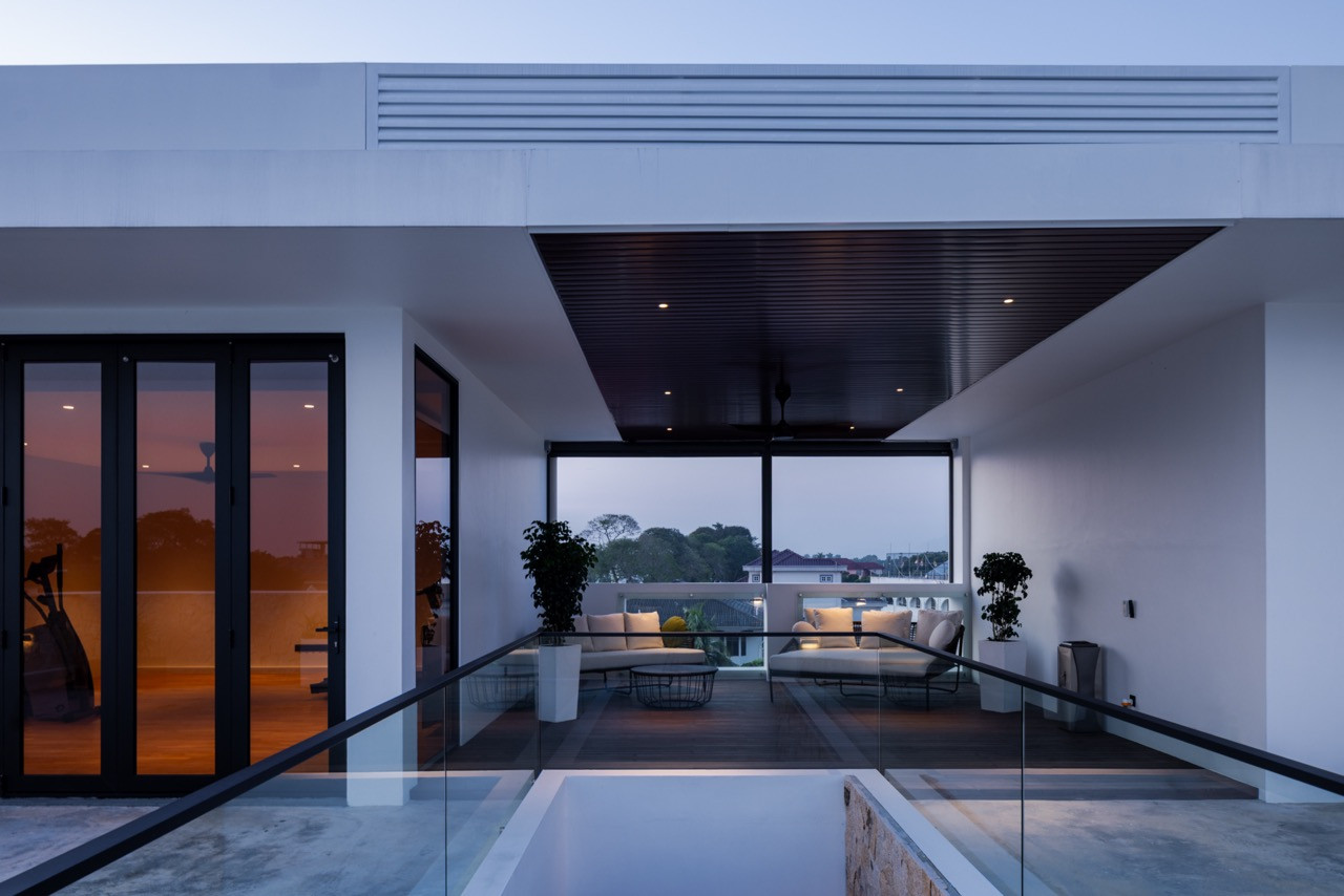 The rear villa’s garden elevation at night, with the koi pond fountain jets lit from below, the frangipani tree spot-lit against the white perimeter wall, and the living room warm behind the glass sliding doors
