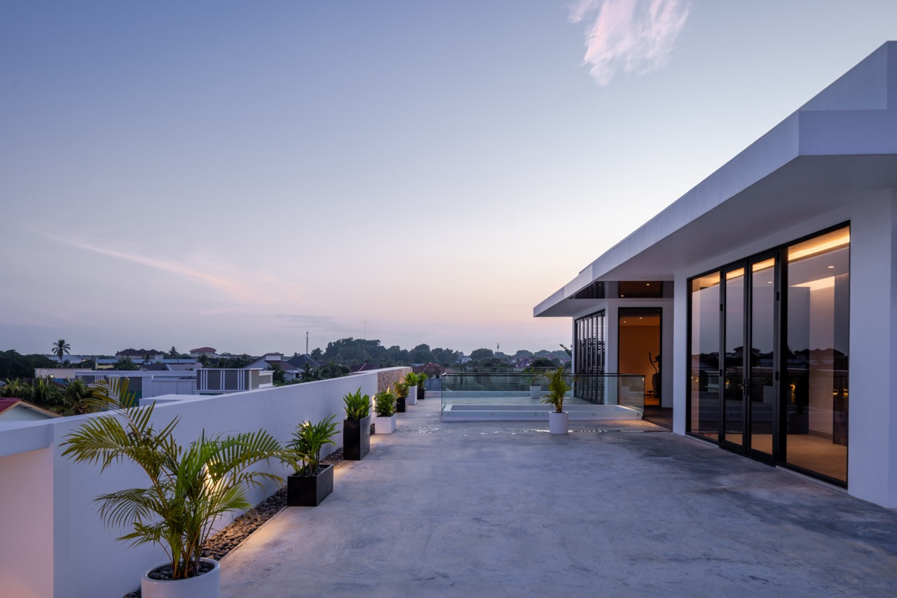 The rear villa at dusk, with the pond fountain jets illuminated and the living room’s cove-lit ceiling and dark grey television wall visible through the fully open bifold entrance