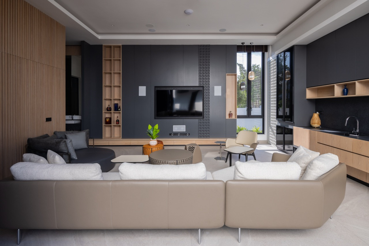The villa’s living room from the interior, with a tan leather corner sofa, dark grey feature wall with geometric relief panelling, open timber shelving, and a bar counter to the right