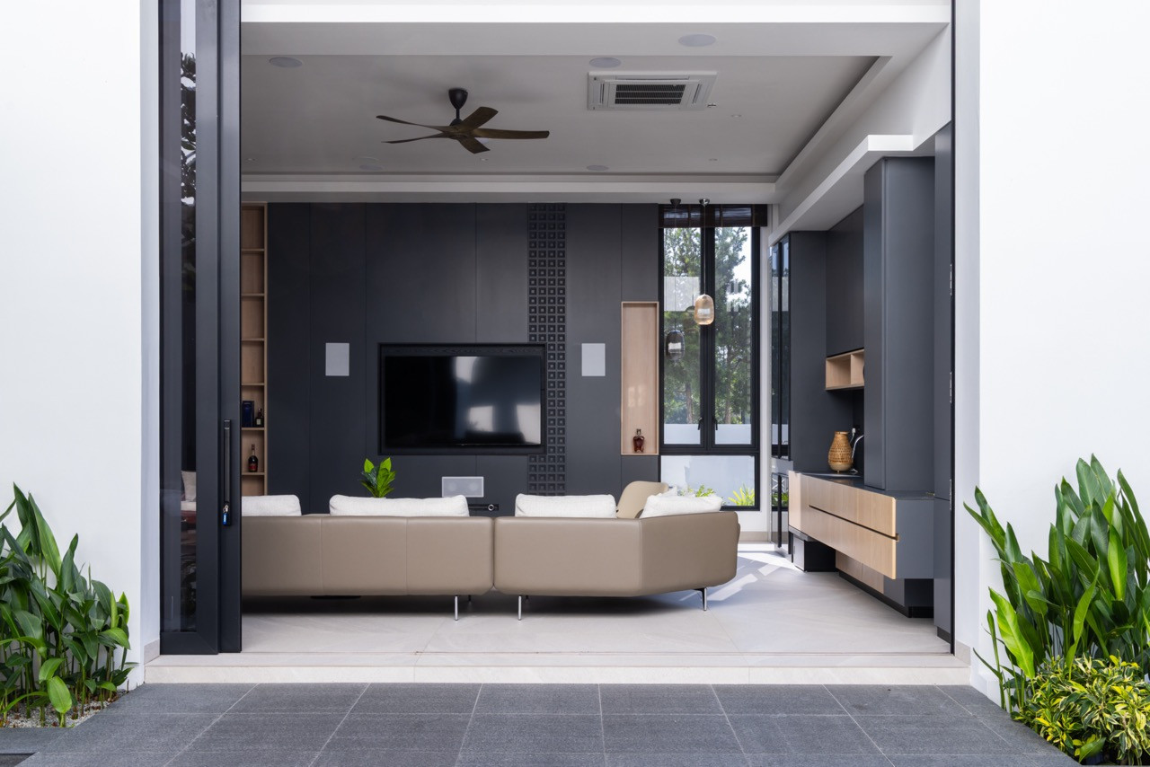 The villa’s living room viewed from the pond terrace with the bifold wall fully open, showing the dark grey television wall with textured panel inserts, open timber shelving, and a kitchen counter to the right