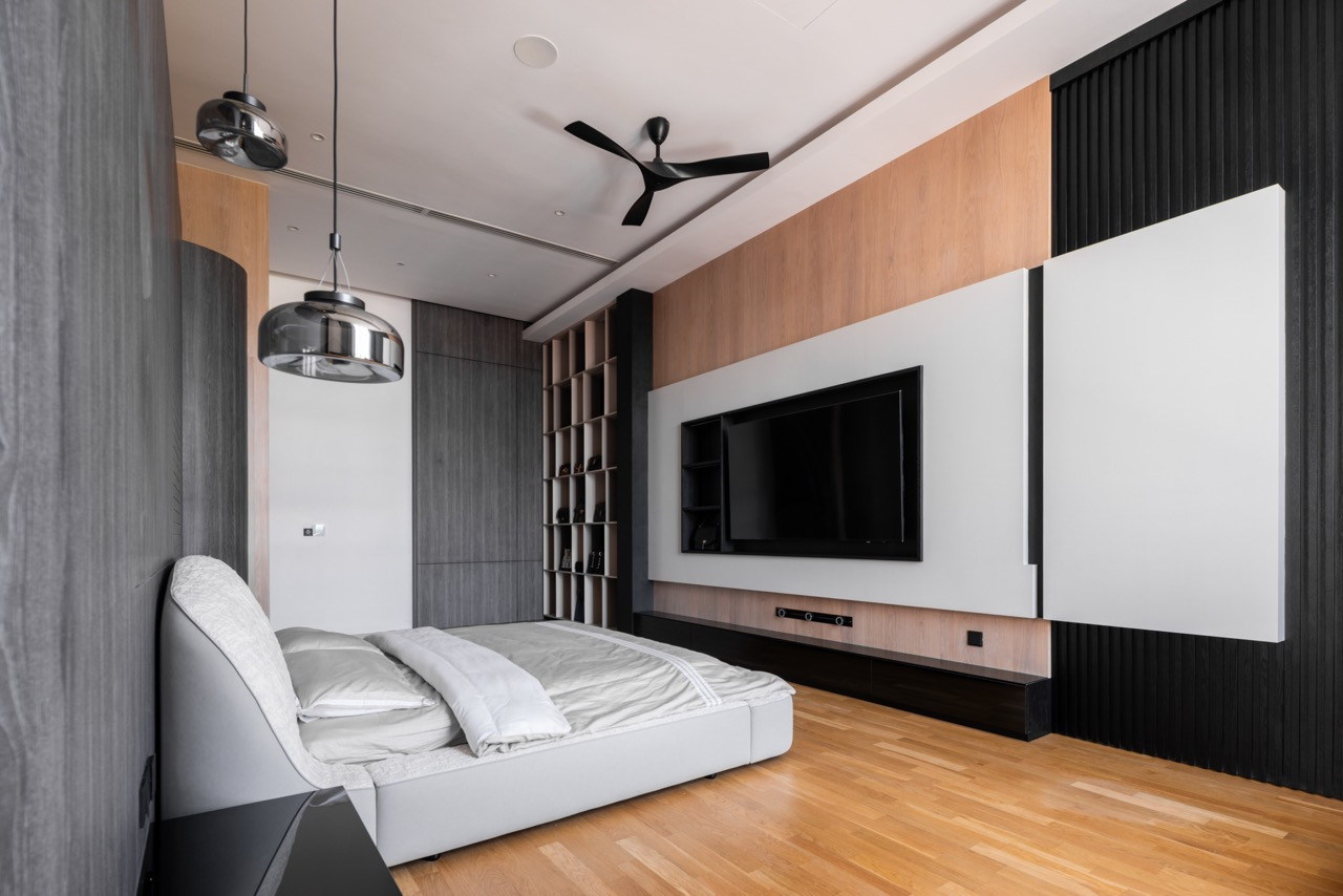 The master bedroom viewed from the sitting area, showing the television wall flanked by dark fluted timber panels, an open shoe cabinet at the rear, and chrome dome pendant lights above