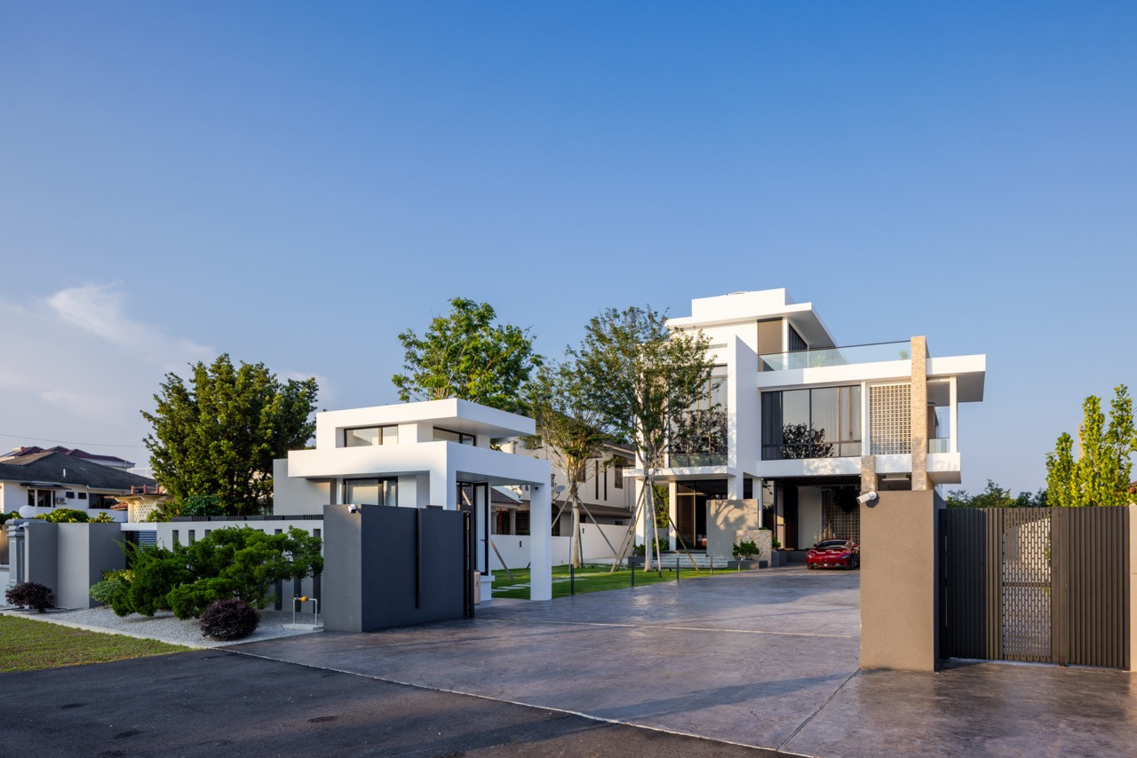 The full street frontage at dawn, showing the main residence at right with its stacked white volumes, stone column, and louvred screens, alongside the guardhouse and perimeter gate at left