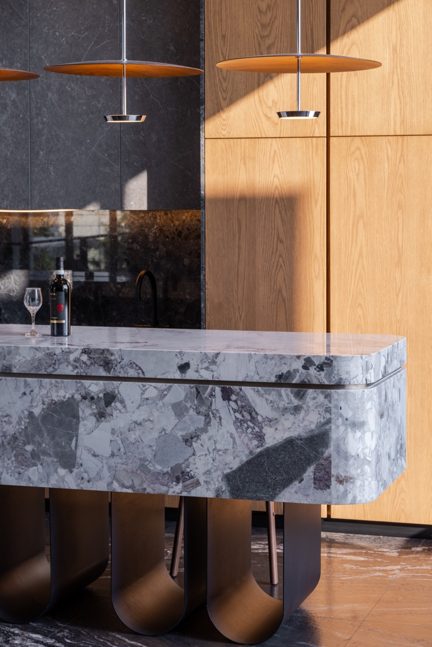 The kitchen island, clad in Ori Grey and Argos Black stone with softened curved edges, beneath a pair of copper-disc pendants. Timber cabinetry