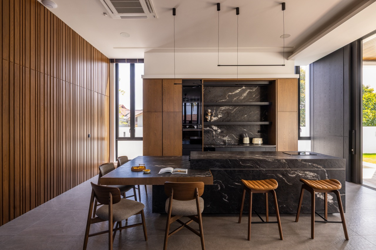 The villa's kitchen and dining area, with a dark stone island and integrated dining table extension, timber-fronted cabinetry, a dark veined stone splashback, and a wall of vertical timber slats to the left