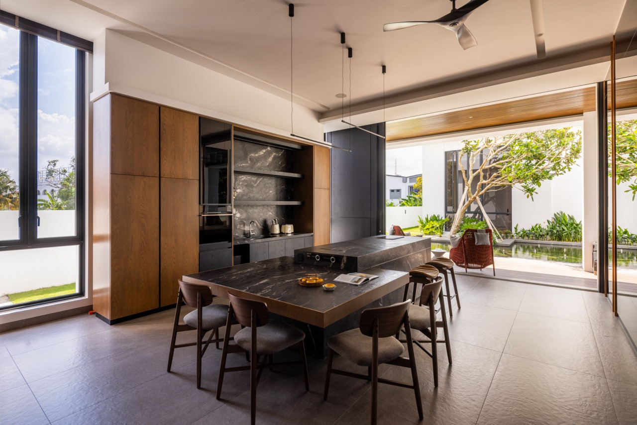 The villa kitchen and dining room in full, with the dark stone dining table and island at centre, timber cabinetry with open stone-clad shelving behind, and the koi pond and garden terrace visible through the open bifold wall to the right