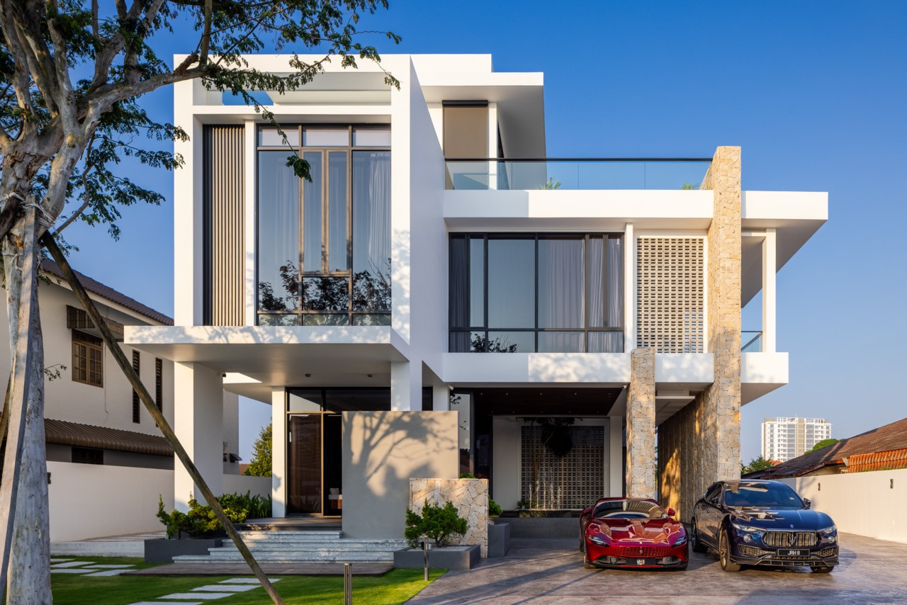 The front elevation of the main residence in morning light, with the stone-clad columns, louvred screen panel, stacked white cantilevers, and glass balustrade at the roof terrace, flanked by the rear villa visible at far left