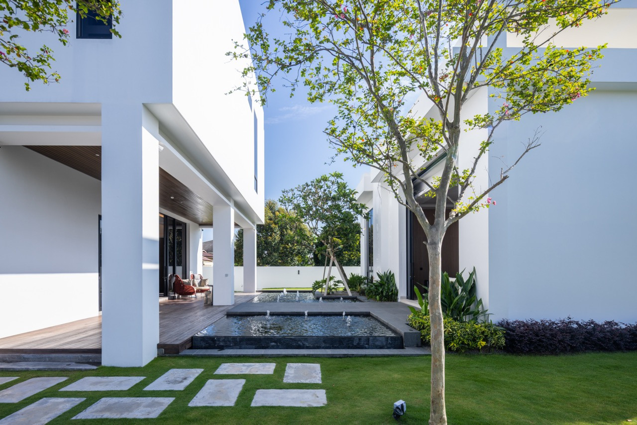 The rear villa seen from across the second koi pond, with a stone-paved path running between the fountain jets toward the open bifold entrance and the single-storey living room beyond