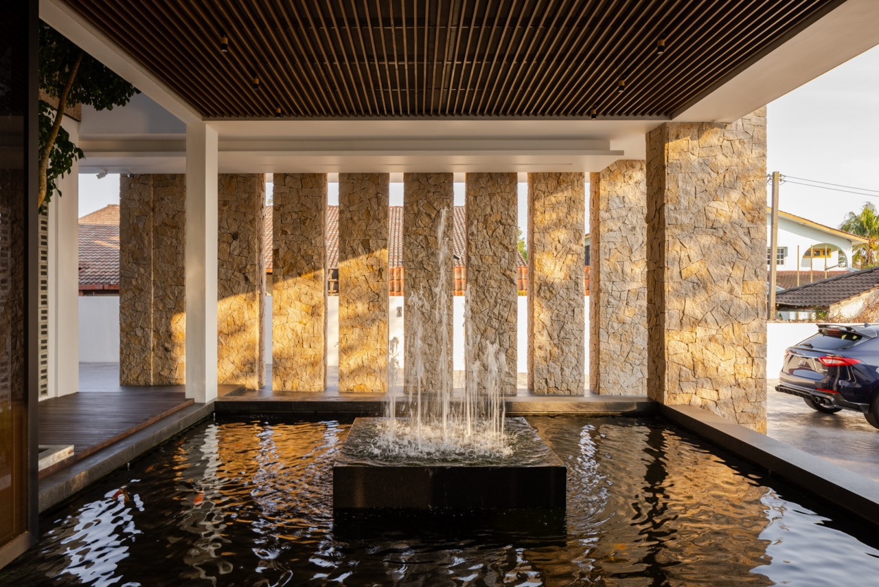 The front entry koi pond and water curtain fountain beneath the main residence's timber-slatted porte-cochère, framed by the rough-cut stone wall