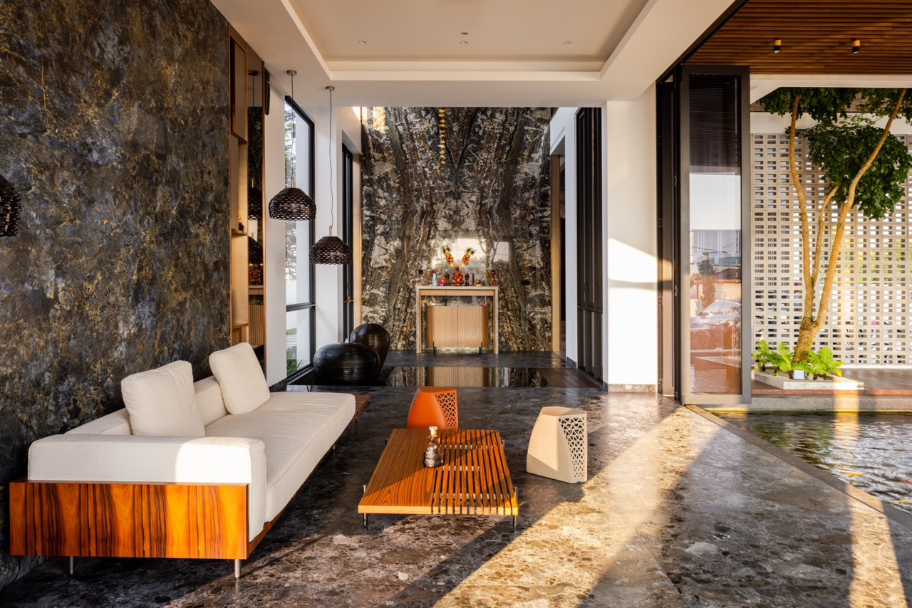 The ground-floor living room of the rear villa, with a white sofa on a zebrawood base, a slatted timber coffee table, and the altar set against a full-height Olive Wave Granite slab at the rear, flanked by the koi pond to the right
