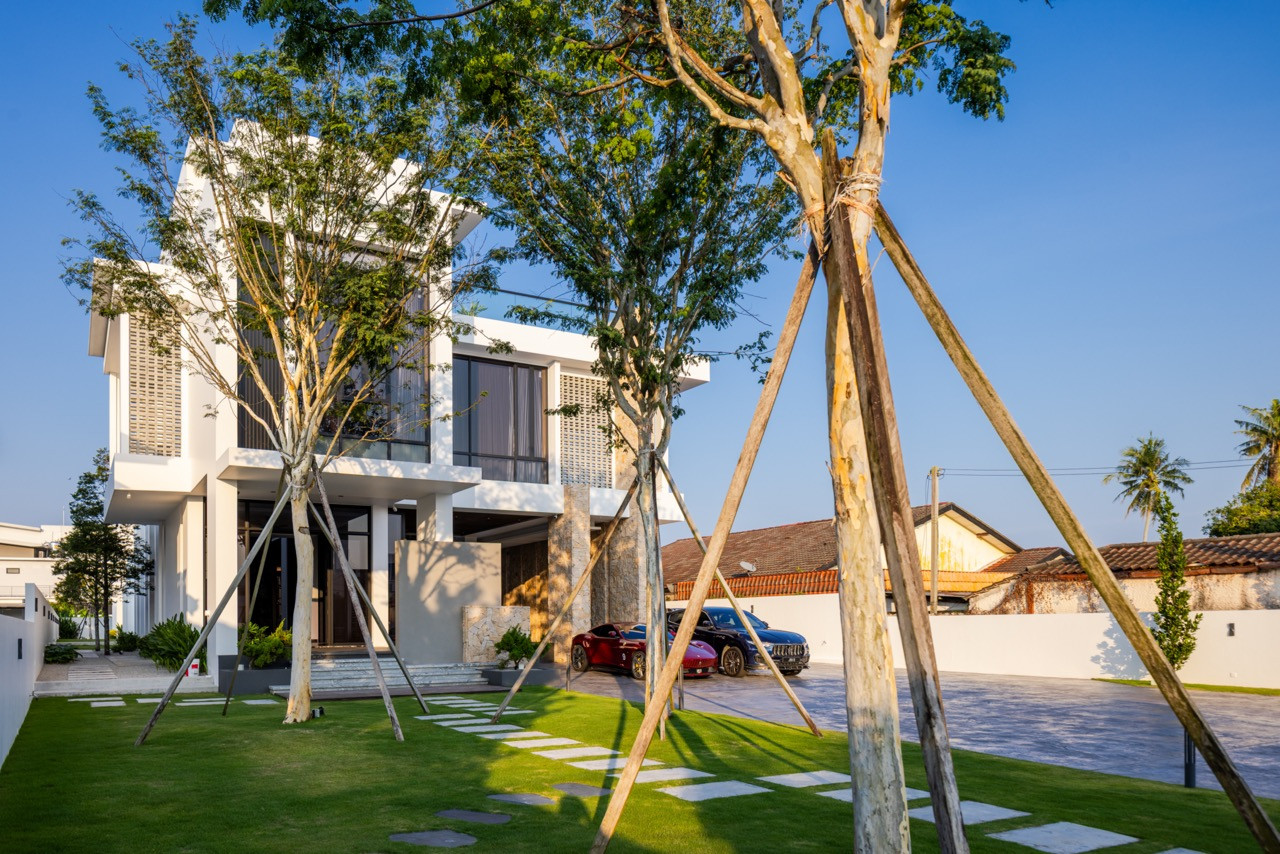The street frontage in full, with the perimeter gate in dark grey and gold-toned vertical-louvred panels, the main residence rising behind the tree planting, and the single-storey rear villa visible to the left