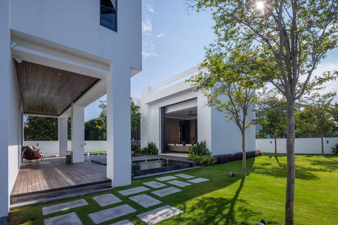 The rear garden and villa from the side, showing the timber-decked terrace to the left, the still-water pond and fountain, stepping stone path across the lawn, and the villa’s open living space beyond