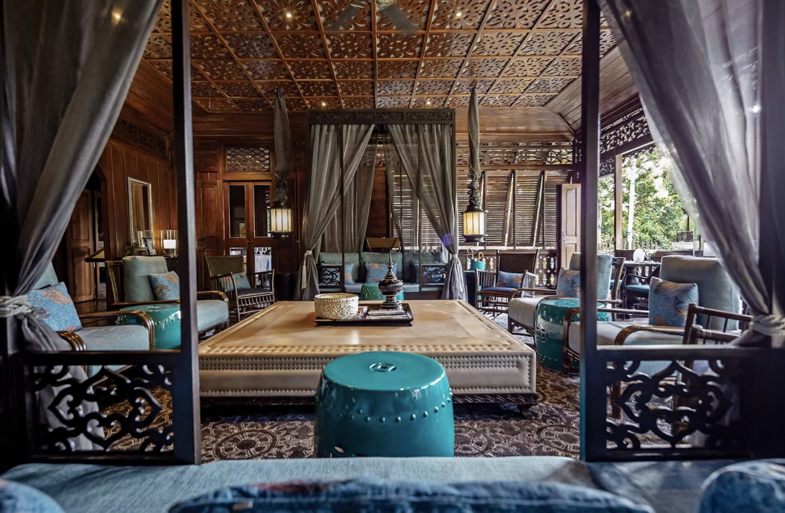 The Parlour Lounge inside the original teak house, with carved fretwork ceiling panels, louvred shutters, and rattan furniture, references the building's late-19th-century colonial history.