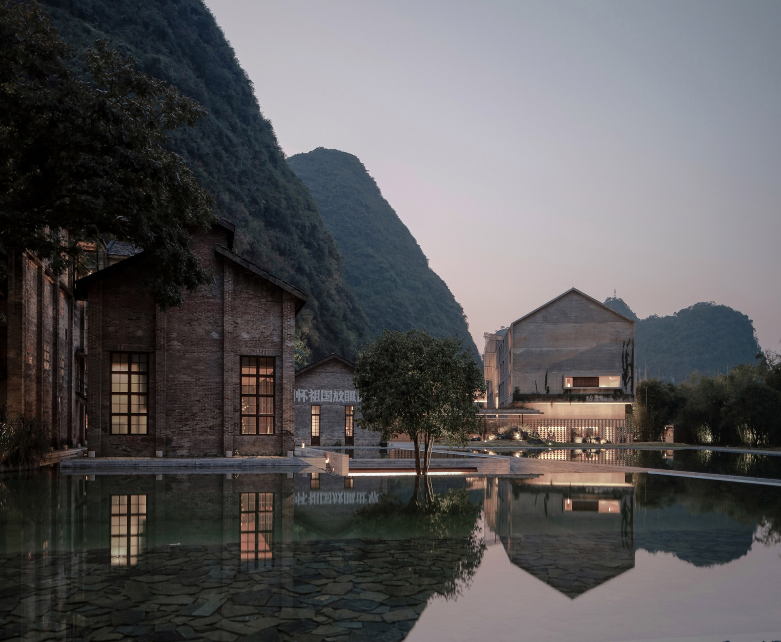 The original 1960s sugar mill buildings reflected in the sunken plaza at dusk, with the karst peaks of Yangshuo rising directly behind the site