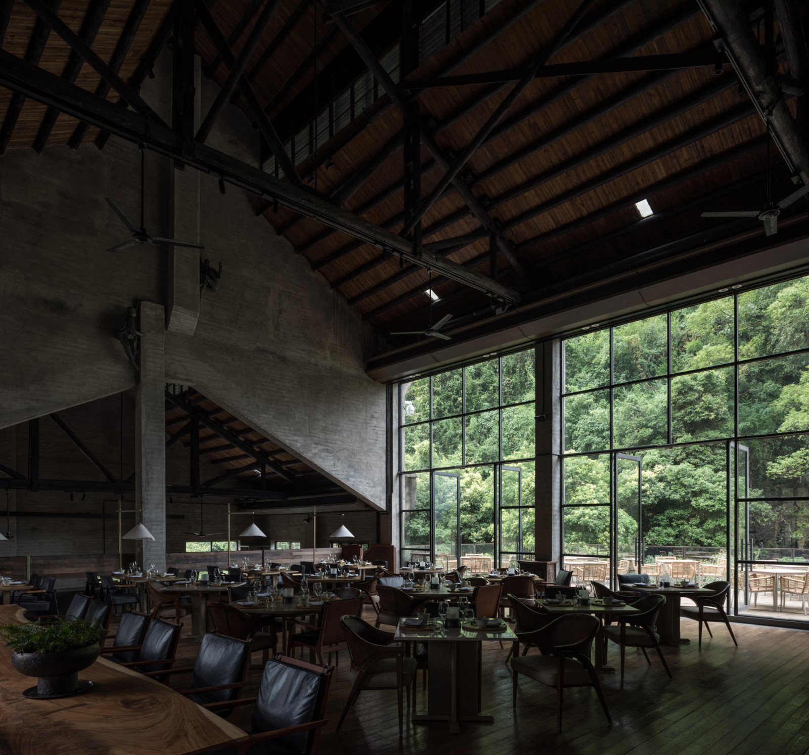 The restaurant inside the original mill building, its industrial roof trusses and concrete columns left exposed; floor-to-ceiling glazing opens the space towards the Li River and surrounding forest