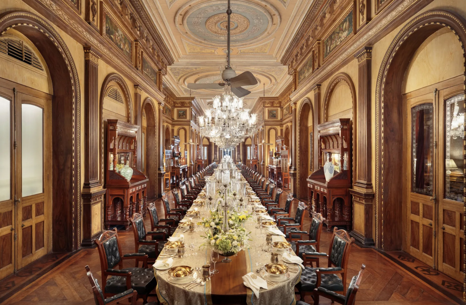 The state dining room, where a single table seats 101 guests beneath painted ceilings and crystal chandeliers. The chairs are carved rosewood with green leather upholstery, original to the palace