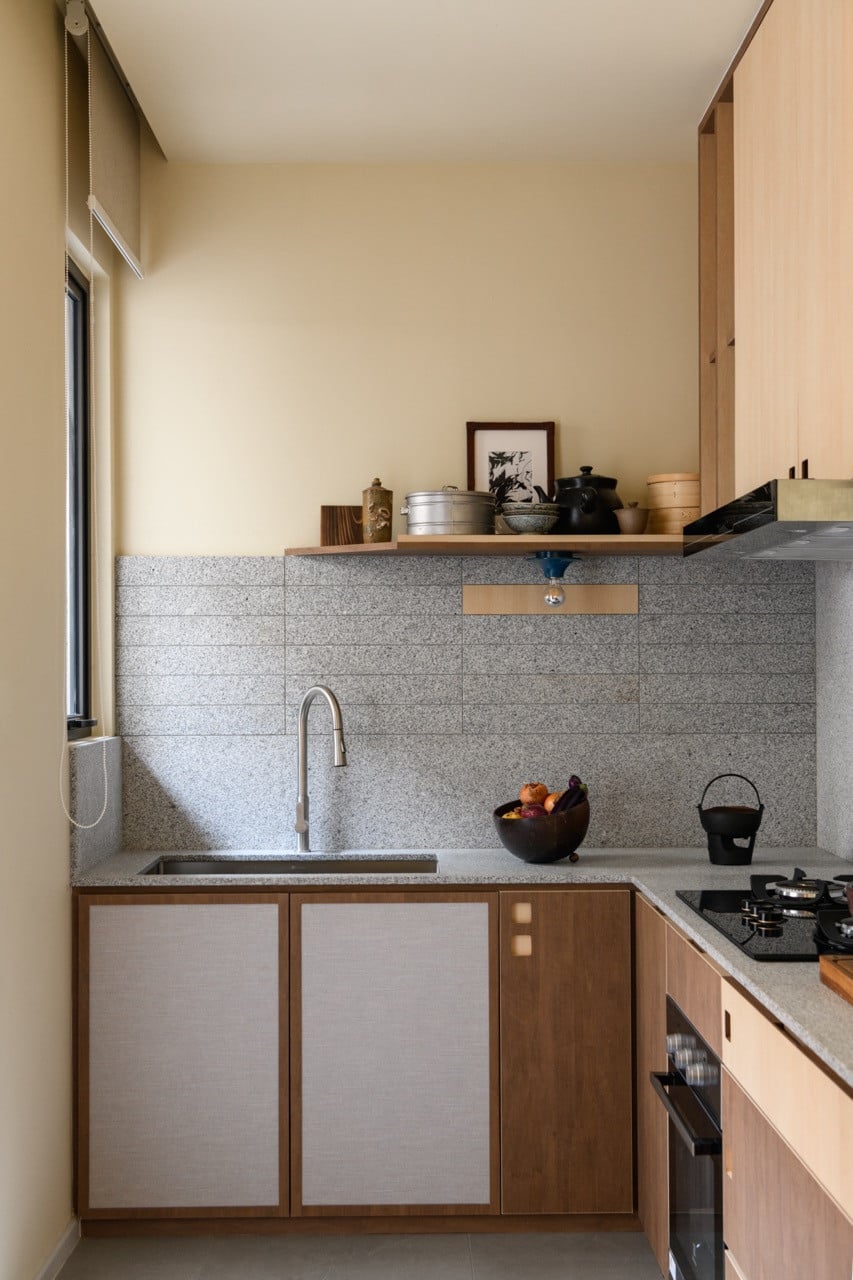 The compact kitchen with a grey granite countertop and matching tiled backsplash, wood cabinetry with fabric inset panels, and an open shelf displaying ceramics