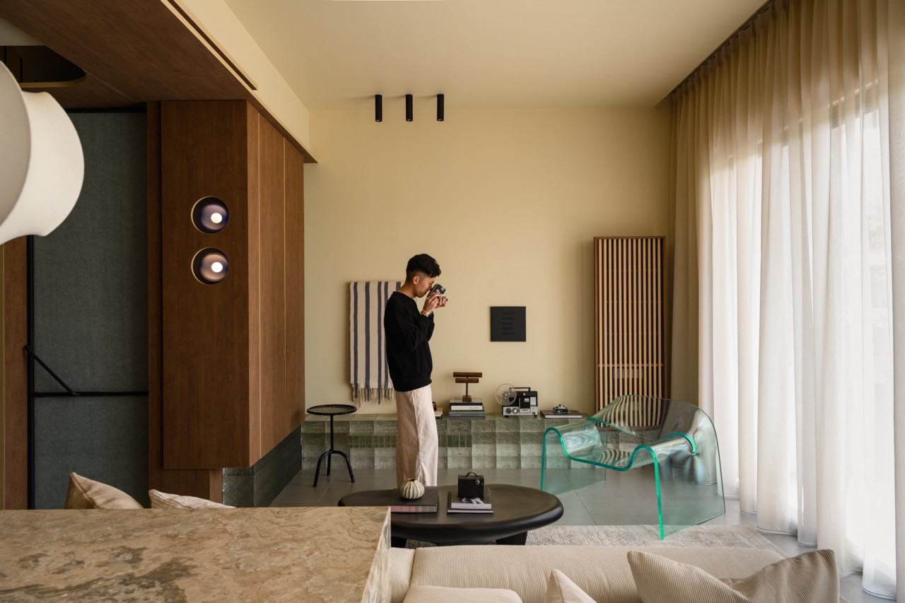 The living room with a raised glass brick platform, an acrylic chair, a vertical wood slat panel, and a pair of circular wall-mounted lights beside the kitchen box volume