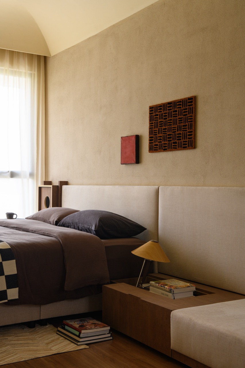 A detail of the master bedroom showing the upholstered headboard, rust-toned bedding, yellow conical bedside lamp, and a vaulted plaster ceiling