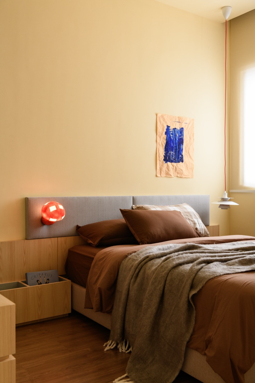 A bedroom with an upholstered grey headboard, rust-toned linen bedding, a red sculptural lamp, and a blue painting on kraft paper pinned to the yellow wall