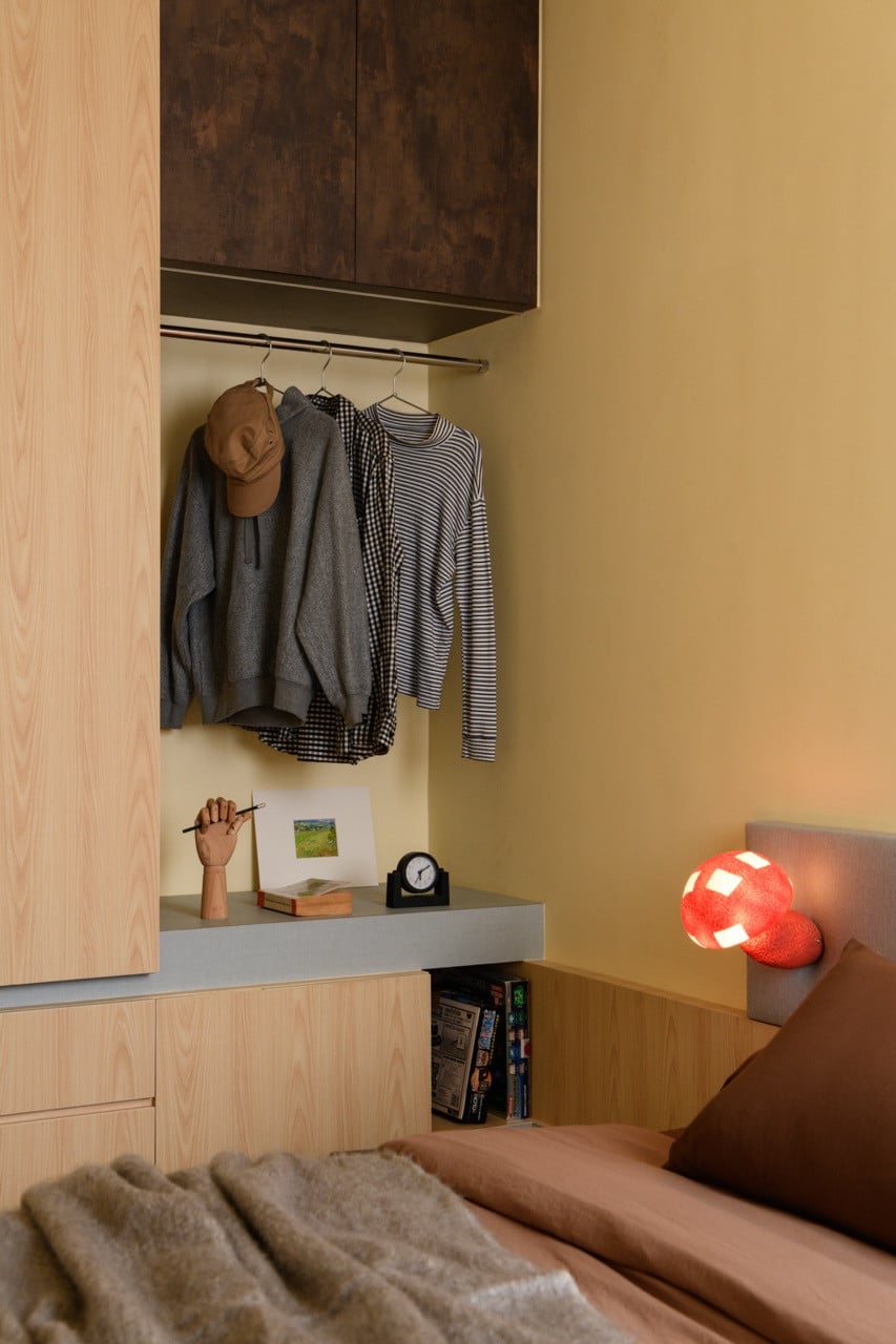 Open wardrobe detail in a bedroom showing light wood cabinetry, a hanging rail beneath dark upper cabinets, and a red sculptural lamp on the bedsi
