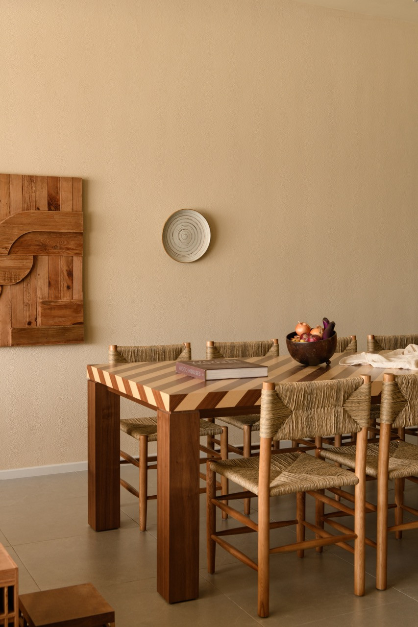 The dining area with a striped two-tone wood table, rush-seat chairs, and a sculptural relief artwork in reclaimed wood on the adjacent wall