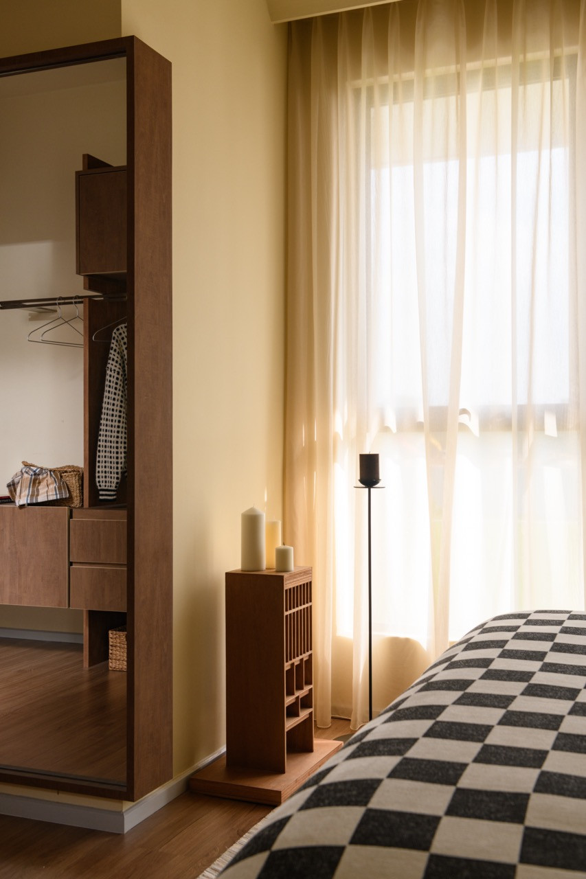 A detail of the master bedroom showing the open wardrobe unit in dark wood beside a slatted bedside cabinet topped with pillar candles, with sheer curtains filtering light behind