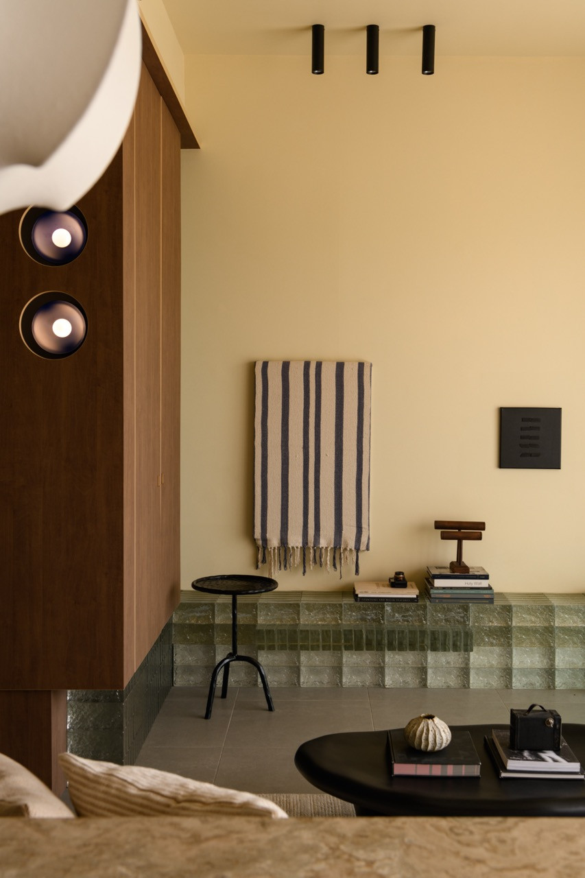 The raised glass brick platform area with a striped fringed textile hung on the wall, a small black side table, a stack of books, and two circular wall lights on the adjacent dark wood panel