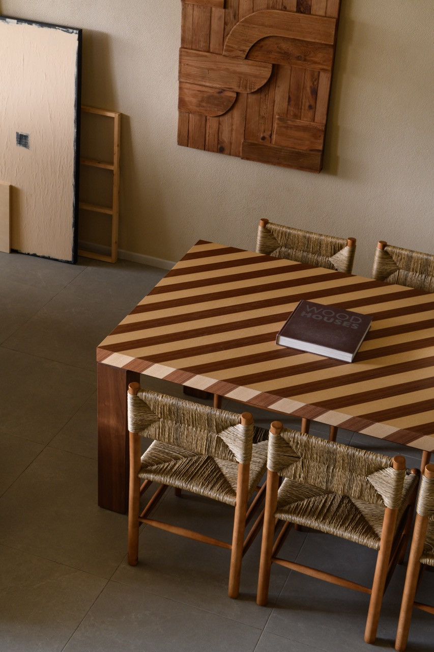 A close-up of the striped two-tone wood dining table top with rush-seat chairs, and a sculptural reclaimed wood wall relief mounted behind