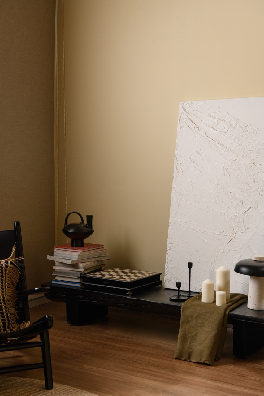 A corner of the living room with a black low bench holding stacked books, a chess set, and a ceramic teapot, with a large textured white canvas leaning against the wall beside pillar candles