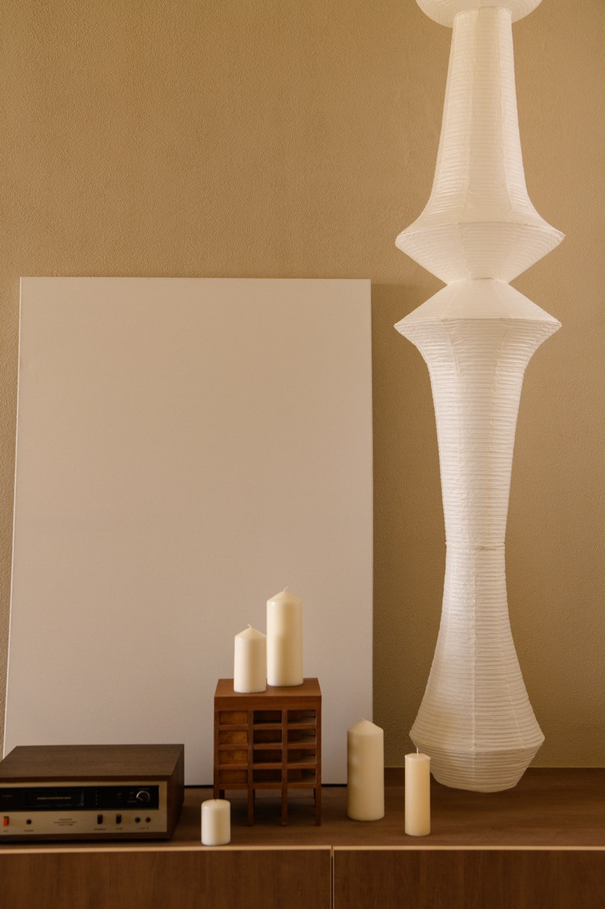 A sideboard vignette in the living area with pillar candles on a small slatted wood stand, a vintage audio receiver, and a tall ribbed paper floor lamp beside a blank canvas