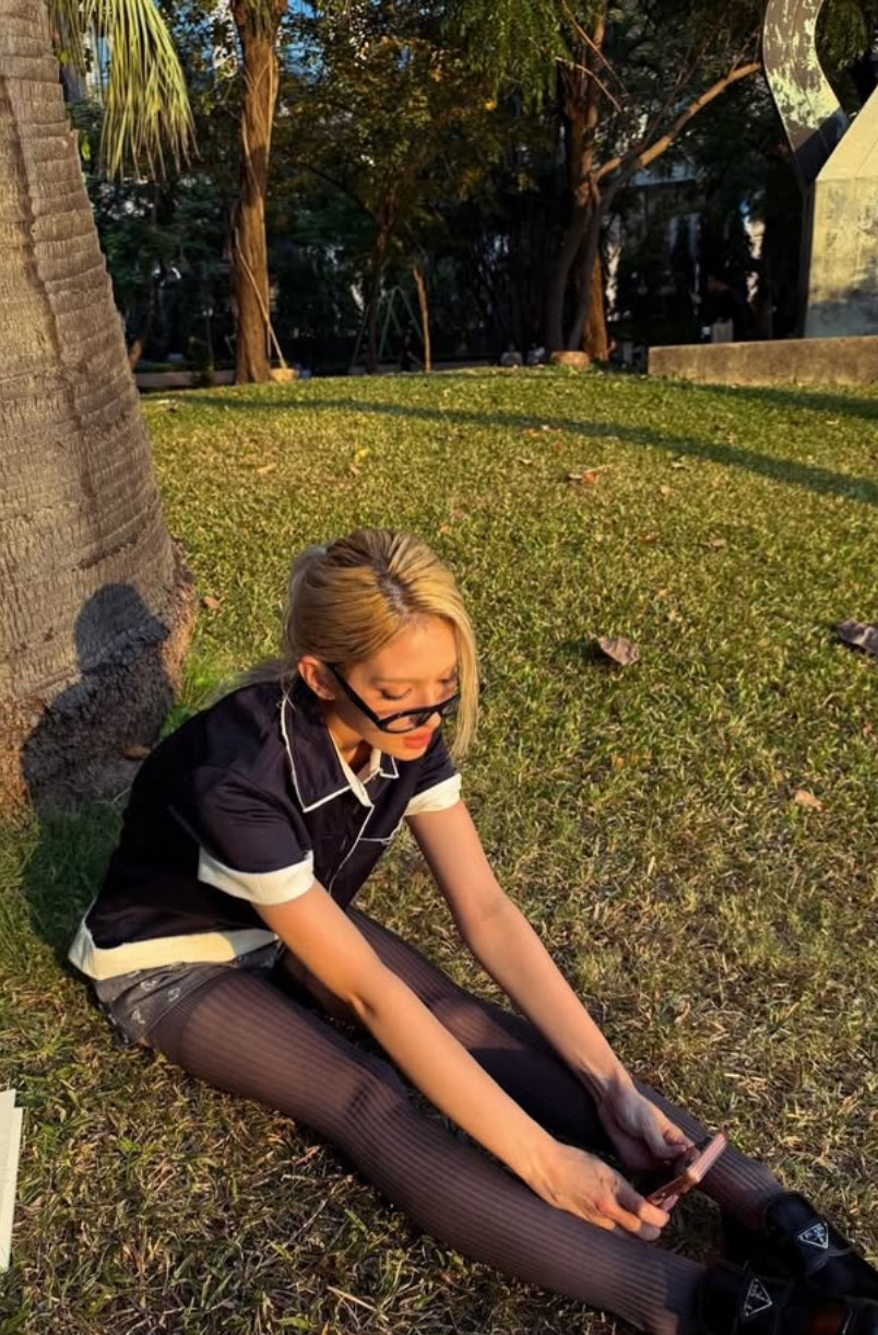 Somi sits on the grass in a navy Prada polo, mini shorts, sheer tights and triangle-logo loafers (Photo: @somsomi0309/Instagram)
