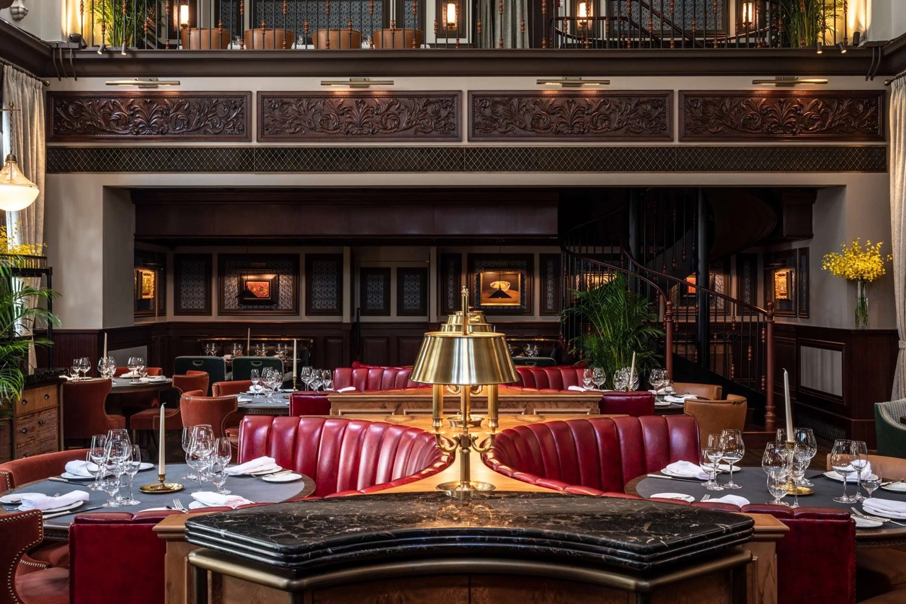 The double-height main dining room of The Magistracy at Tai Kwun, designed by Joyce Wang Studio, with the original carved timber frieze and mezzanine gallery of the former Central Magistracy courtroom visible above the red leather banquettes