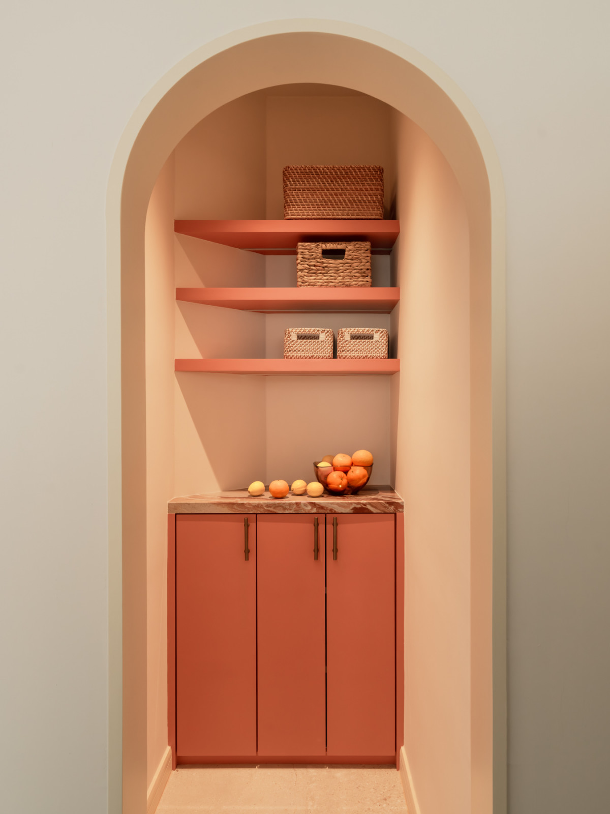 A kitchen pantry nook framed by a full plaster arch, with terracotta-pink open shelving, a marble countertop, and woven baskets throughout