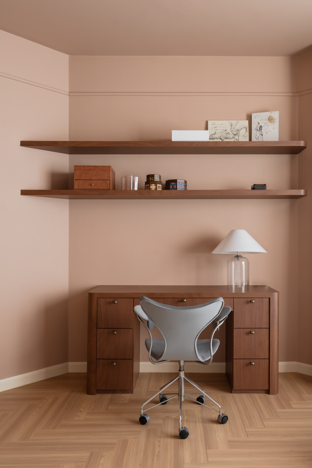 A study corner with walnut floating shelves, a dark timber pedestal desk, and a chrome swivel chair on herringbone timber flooring