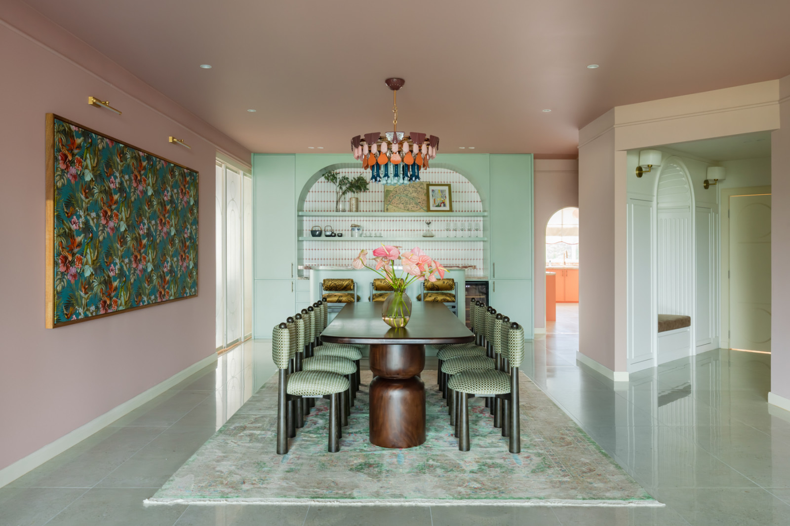 The dining room viewed head-on, with the tropical botanical artwork on the left wall, the Lladró chandelier overhead, the Dad’s Woods dining table at centre, and the mint-green bar and arched nook beyond