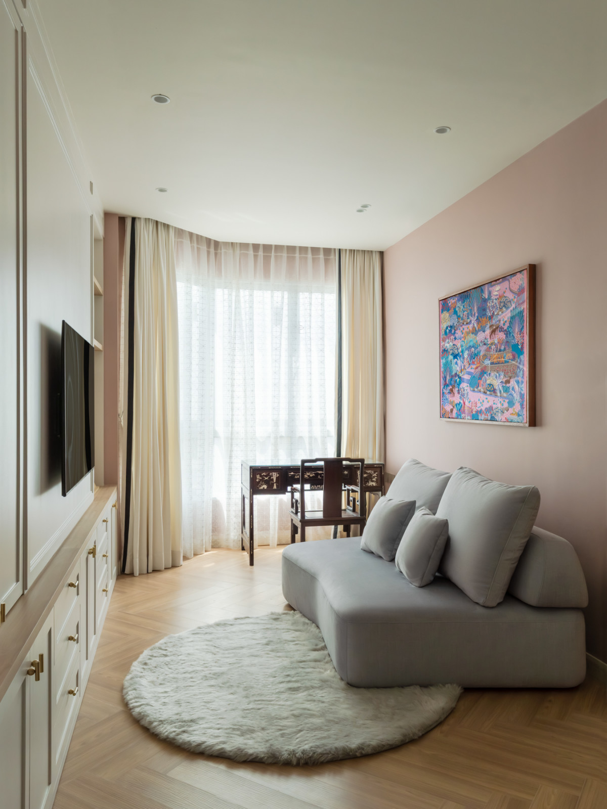 The family room from the opposite angle, with a rounded grey sofa on a circular shag rug, built-in white cabinetry with brass hardware, and floor-length sheer curtains