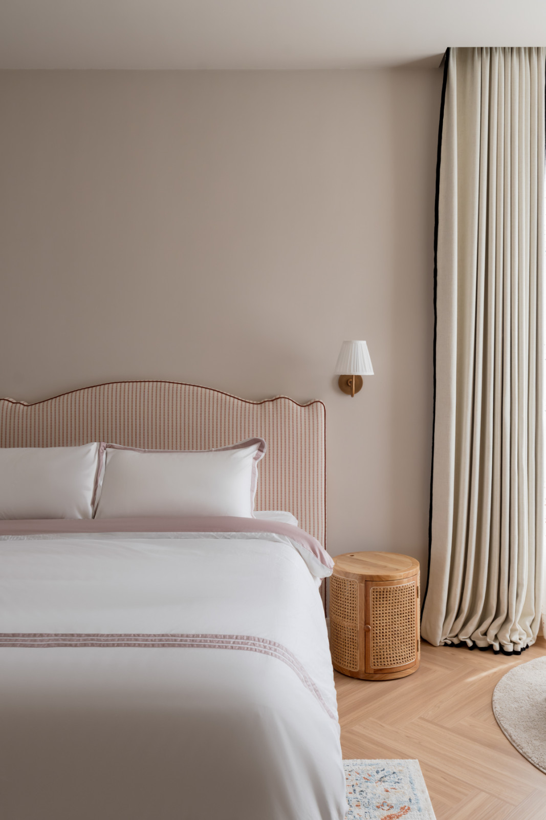 A guest bedroom with a scallop-edged headboard upholstered in a pink striped fabric, a cylindrical rattan bedside table, and floor-length curtains with a navy trim