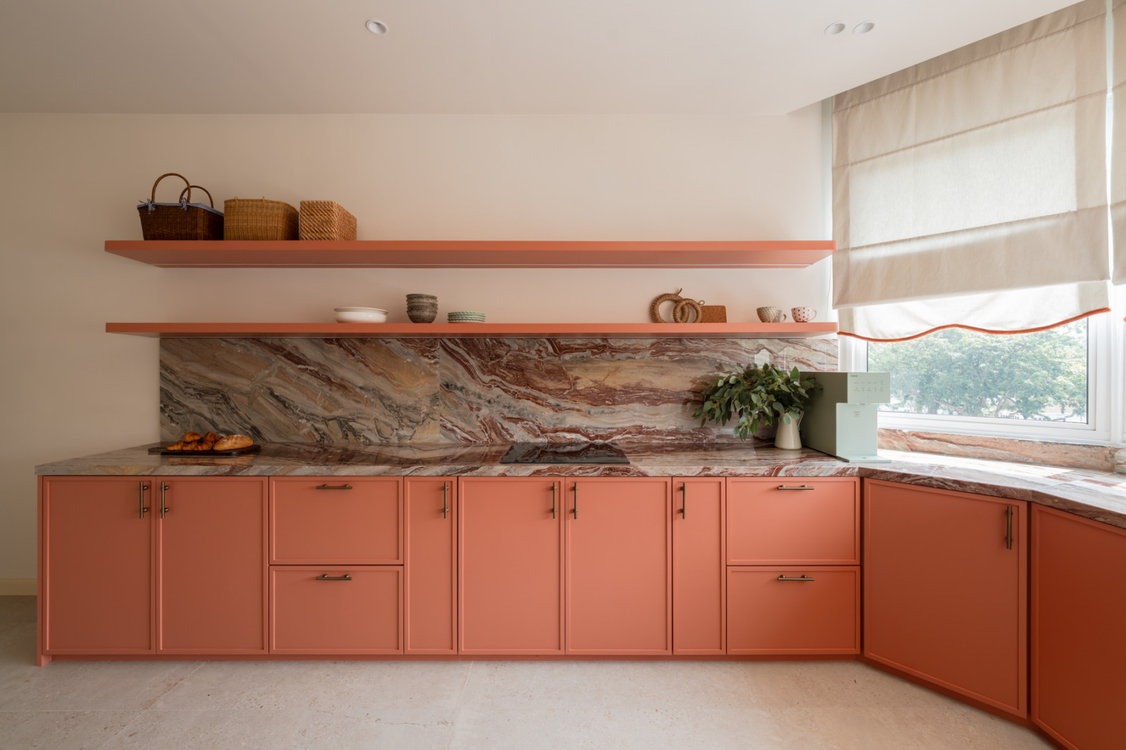 The kitchen cabinetry wall, with a full slab of red and grey veined marble as the backsplash and terracotta-pink lower cabinets below matching open shelving