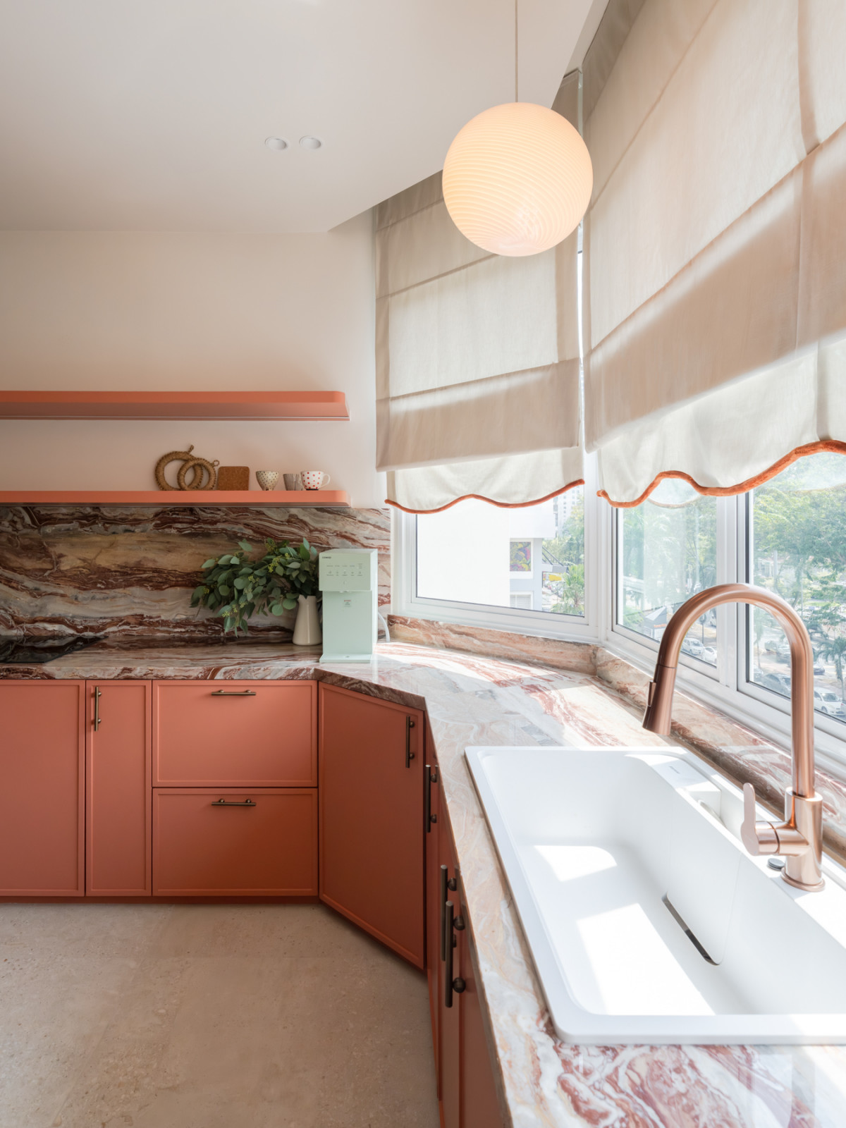The kitchen sink corner, where the veined marble countertop wraps around the angle, a globe pendant hangs overhead, and linen blinds with coral scalloped edging frame the city view