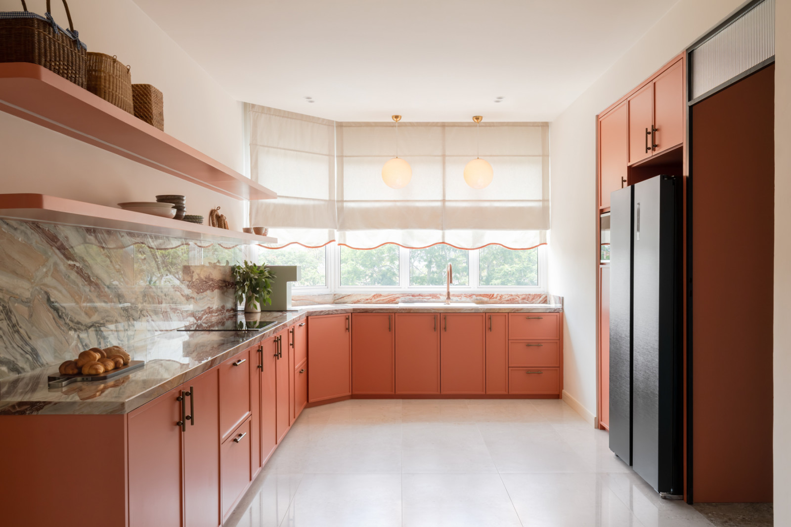 The kitchen, with terracotta-pink cabinetry running the full length of the L-shaped layout, a veined marble backsplash, and linen roman blinds with a coral scalloped trim at the windows
